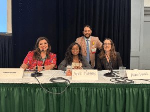 Four people sit at a panel table with name cards reading Monica N. Sauceda, Irene Musambi, and Amber Weeks; a man stands behind them. They are indoors with a green and white tablecloth.